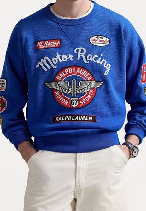 Homme portant un sweat-shirt bleu Ralph Lauren Motor Racing avec des patchs brodés et un pantalon blanc, les mains dans les poches.