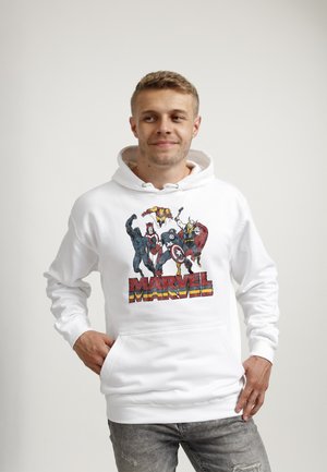 Młody mężczyzna noszący białą bluzę z kapturem Marvel z postaciami Black Panther, Scarlet Witch, Iron Man, Captain America i Thor, stojący z ręką w kieszeni.