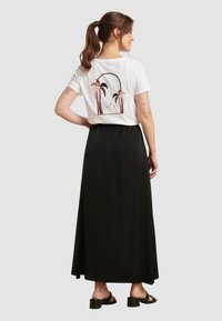 Witte t-shirt met palmboomgrafiek en "Lost In" tekst op de achterkant, gecombineerd met een lange zwarte rok. Model draagt zwarte sandalen.