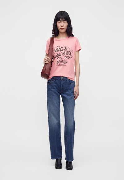 Γυναίκα που φοράει ροζ γραφικό T-shirt, μπλε τζιν, μαύρα μποτάκια στον αστράγαλο και καφέ τσάντα ώμου σε μονόχρωμο ανοιχτό φόντο.