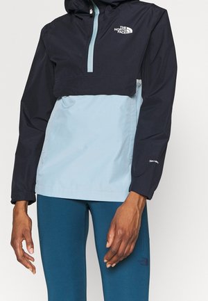 Osoba ubrana w dwu kolory kurtkę North Face w czerni i jasnym niebieskim z kapturem oraz niebieskie legginsy, stojąca na jednolitym tle.