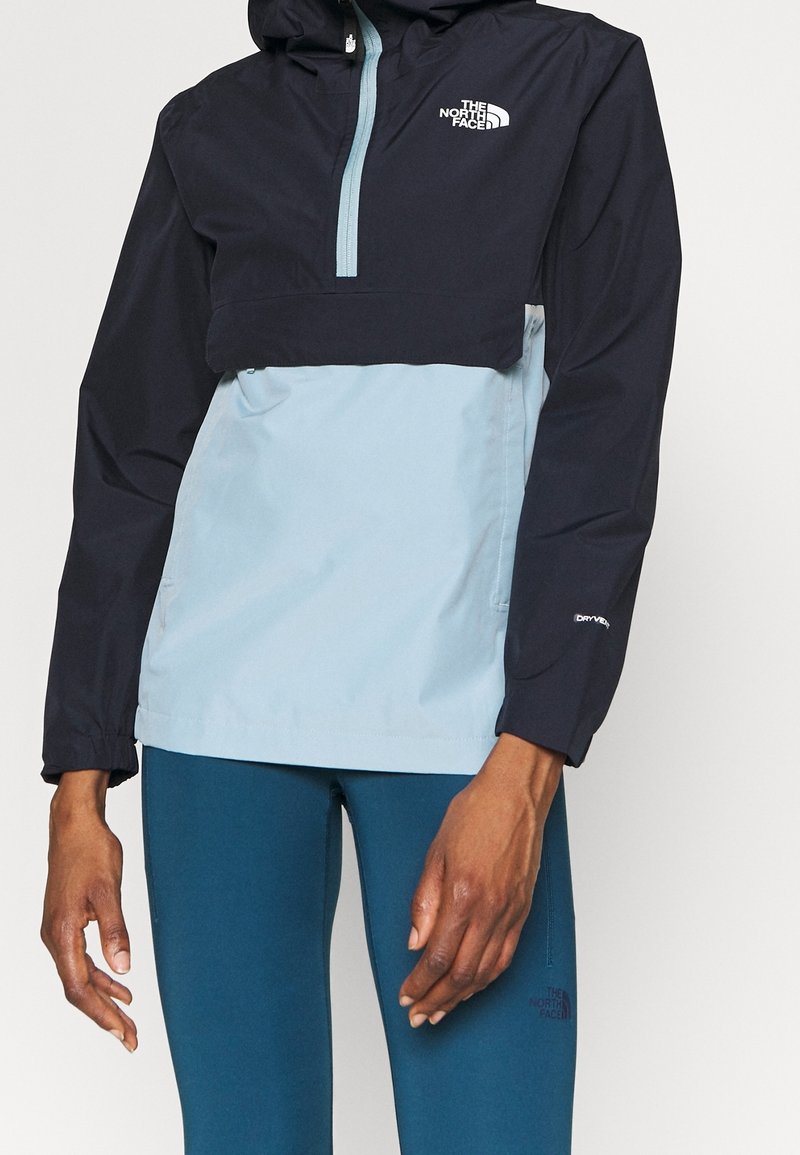 Personne portant une veste North Face bicolore noir et bleu clair avec capuche et un legging bleu, debout devant un fond uni.