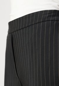 Melnas pinstripe bikses ar gludu tekstūru, kas aprīkotas ar platu jostu un diskretām brūnganām svītrām, kas vertikāli iet pa audumu.
