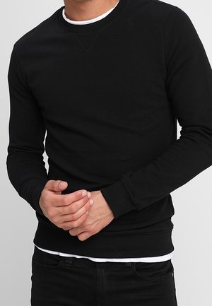 Hombre vestido con un suéter ajustado de manga larga negro sobre una camiseta blanca, con las manos entrelazadas a la altura de la cintura, sobre un fondo liso de color gris claro.