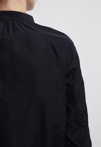 Marinblå, lätt, texturerad bomberjacka med ribbad krage, med en slät yta och rynkade detaljer på baksidan.