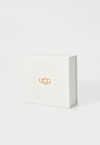 Bijela kutija od kartona s narančastim UGG logotipom u središtu, okružena uzorkom sunčevih zraka.