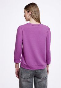 Felpa viola con maniche a tre quarti, collo a costine, texture morbida e orlo aderente, abbinata a pantaloni in denim grigi.