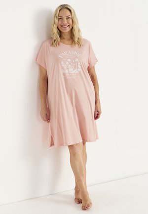 Kobieta uśmiechająca się, stojąca boso przy białej ścianie, ubrana w luźną różową sukienkę do kolan z tropikalnym nadrukiem "Paradise Islands".