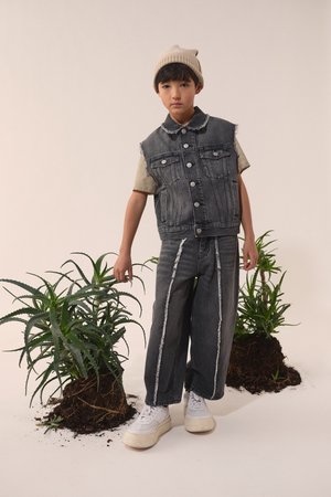 Jeansväst och vida byxor i mörkgrått med fransade kanter och vit sömnad. Modellen bär en beige mössa och vita sneakers. Gröna växter i bakgrunden.