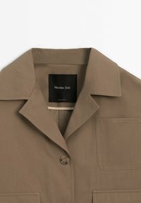 Veste beige à col avec un bouton, poches plaquées sur la poitrine et en bas, et une étiquette noire portant l'inscription « Massimo Dutti » à l'intérieur du col.