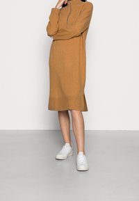 Stickad brun klänning med hög krage och halv dragkedja. Har långa ärmar och sidofrånskärningar i fållen, i kombination med vita sneakers.