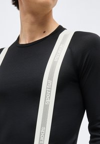 Άτομο που φοράει μαύρο μακρυμάνικο πουκάμισο με υφή και λευκές κάθετες τιράντες με διάτρητο κείμενο.