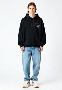 Felpa nera con cappuccio e tasca frontale, caratterizzata da un cuore rosso e una grafica. Abbinata a jeans larghi di colore blu chiaro e sneakers multicolori.