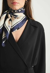Foulard en soie navy et crème, avec des rayures diagonales et des motifs géométriques, noué autour du cou. Blazer noir avec un col pointu.