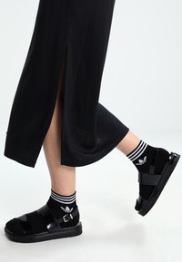 Svarta platåsandaler med glänsande yta och spänne, bärs med svartvita randiga strumpor med en logotyp.