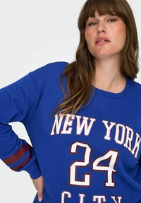 Plava majica s kapuljačom sa natpisom "NEW YORK CITY 24" u bijeloj i crvenoj boji. Ima prugaste crvene i plave detalje na rukavima. Ležeran kroj, teksturirani materijal.
