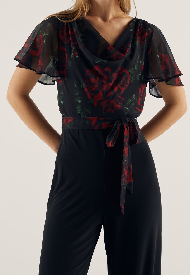 Combinaison noire avec un voile floral transparent orné de roses rouges et de feuilles vertes. Elle possède des manches courtes évasées et une ceinture nouée.