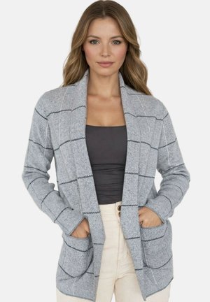 Femme portant un cardigan gris clair à ouverture frontale avec de grandes poches et un motif à carreaux par-dessus un haut gris foncé et un pantalon clair.