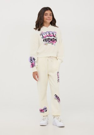 Completo felpa e pantaloni della tuta color crema con testo e grafiche colorate. Il materiale sembra morbido con una vestibilità comoda. Scarpe da ginnastica bianche completano il look.