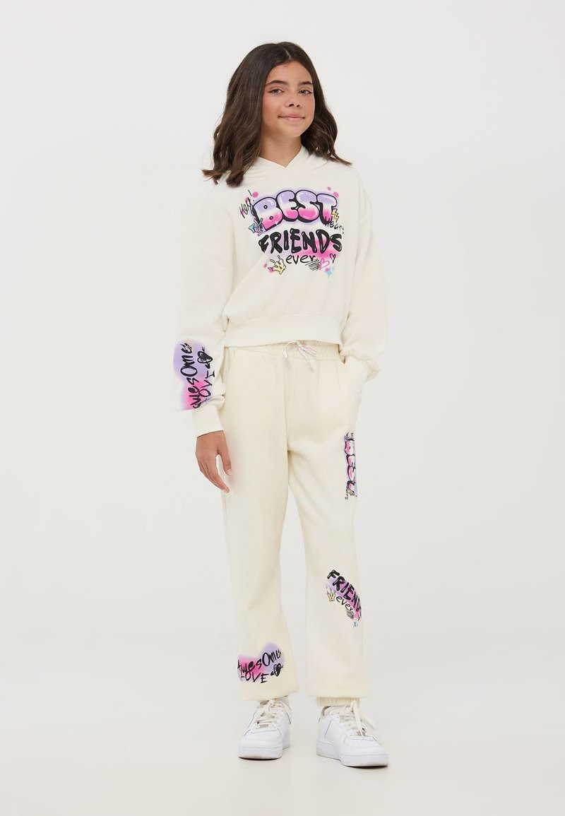 Completo felpa e pantaloni della tuta color crema con testo e grafiche colorate. Il materiale sembra morbido con una vestibilità comoda. Scarpe da ginnastica bianche completano il look.