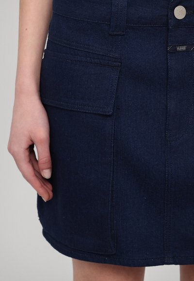 Denimsfarget skjørt i mørkeblå med en jevn tekstur, har to frontlommer og synlig knappelukking. Sømmen er litt buet.