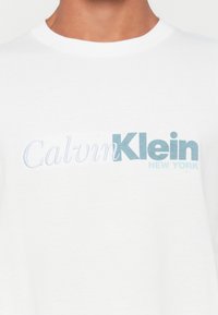 Hvid T-shirt med "Calvin Klein New York" logo i lyseblå og hvide bogstaver på brystet, båret af en person.
