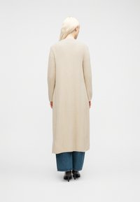 Άτομο με μακριά ξανθά μαλλιά, φορούν μπεζ μακρύ ριγωτό ζακέτα, φαρδιά μπλε τζιν παντελόνια και μαύρα μποτάκια με τακούνι, γυρισμένο προς τα πίσω.