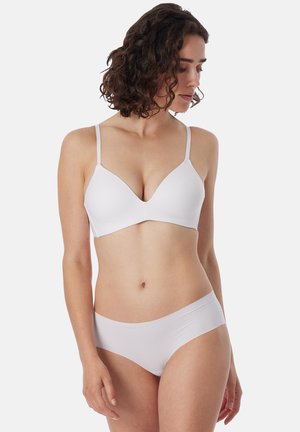 Femme aux cheveux bouclés portant un soutien-gorge blanc sans couture et un sous-vêtement assorti, regardant vers le bas sur sa gauche sur un fond uni.