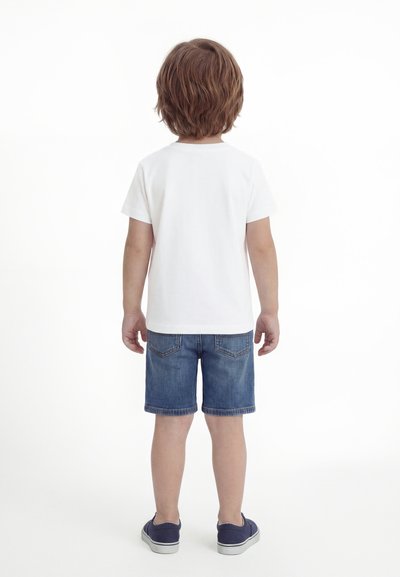 Keskmise pikkusega pruunide juustega laps seisab seljaga vaataja poole, kandes lihtsat valget t-särki, teksapükse ja tumesiniseid spordijalatseid valgel taustal.