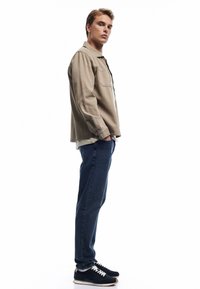 Beige overhemd met knopen en een zak, gedragen over een grijs T-shirt, gecombineerd met donkerblauwe jeans en zwarte sneakers met witte veters.