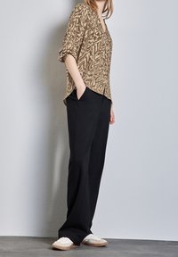 Personne debout avec les mains dans les poches, portant une chemise à manches longues beige à motifs, un pantalon noir à jambes larges et des baskets blanches, devant un mur uni.