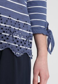 Blå och vit randig topp med spetsdetaljer och ogenomskinliga hål samt paljetter. Rullade ärmar med knytdetalj. Blandad bomullstextil.