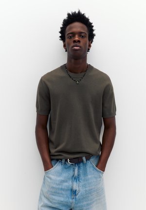 Ung mand med kort krøllet hår, iført en olivengrøn kortærmet skjorte, lyseblå jeans, sort bælte og en perlekæde, hænderne i lommerne.