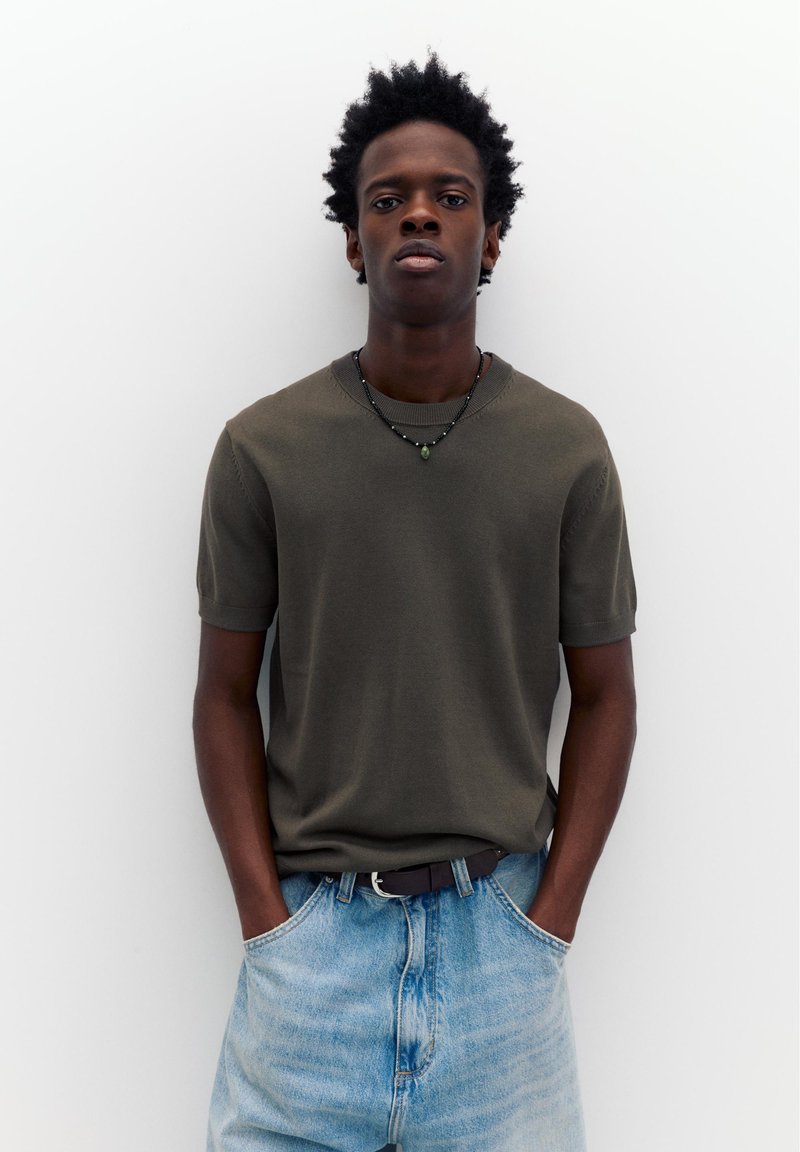 Jeune homme aux cheveux courts et bouclés portant une chemise à manches courtes vert olive, un jean bleu clair, une ceinture noire et un collier de perles, les mains dans les poches.