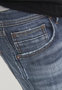 Primer plano del bolsillo deshilachado de unos vaqueros de mezclilla azul con remaches de cobre y costuras amarillas, usados con una camiseta negra.