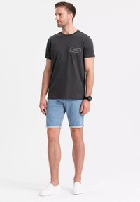 Camiseta de algodón gris oscuro con cuello redondo y bolsillo en el pecho, combinada con pantalones cortos de mezclilla azul claro, con puños doblados, y zapatillas blancas sin cordones.