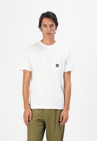 Young man wearing a plain white short-sleeve T-shirt with a small black logo patch on the chest pocket and olive green pants.