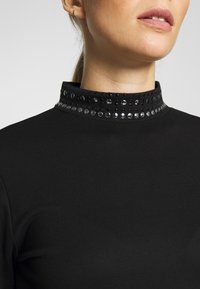 Svart figurnära topp med hög krage som har runda metalliska detaljer och en slät yta. Närbild som framhäver kragedetaljen.