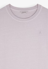 Camiseta de cuello redondo color púrpura claro con un sutil bordado de calavera y tibias cruzadas en el pecho y una etiqueta de la marca Scalpers en el interior del cuello.