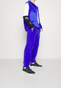 Blå fleeceväst med en ficka över en vit och svart långärmad skjorta. Matchande blå fleecebyxor och svarta sneakers med gula detaljer.