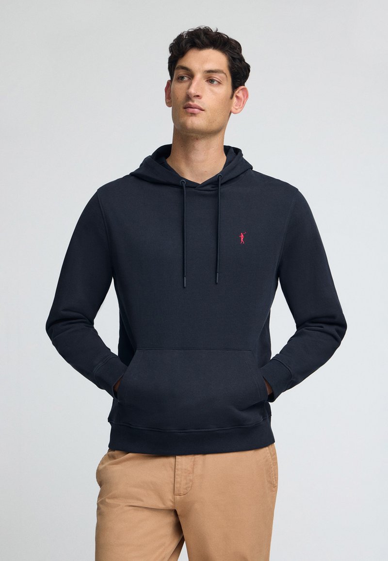 Marinblå hoodie med en känguruficka, dragsko kapuschong och en liten röd logotyp på bröstet. Slät yta, ribbade ärmslut och midja.