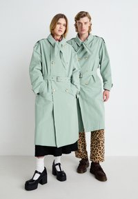 Trench coats verde menta claros com botões duplos. Um modelo usa sapatos pretos de plataforma, o outro está com calças de padrão de leopardo.