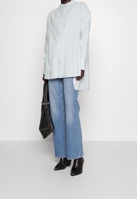 Osoba v nadměrné světle modré pruhované košili, širokých světle modrých džínách, černých špičatých botách a držící volnou černou kabelku.