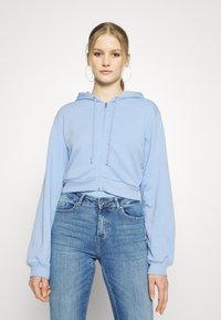 Hoodie cropped de cor azul clara com fecho na frente, cordões e mangas longas, combinado com jeans azuis ajustados. Design simples e textura suave.