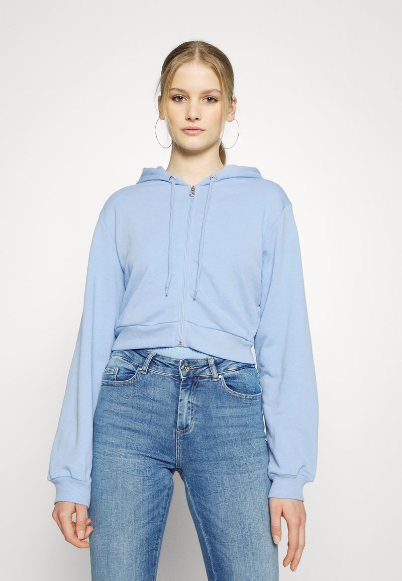 Hoodie cropped de cor azul clara com fecho na frente, cordões e mangas longas, combinado com jeans azuis ajustados. Design simples e textura suave.