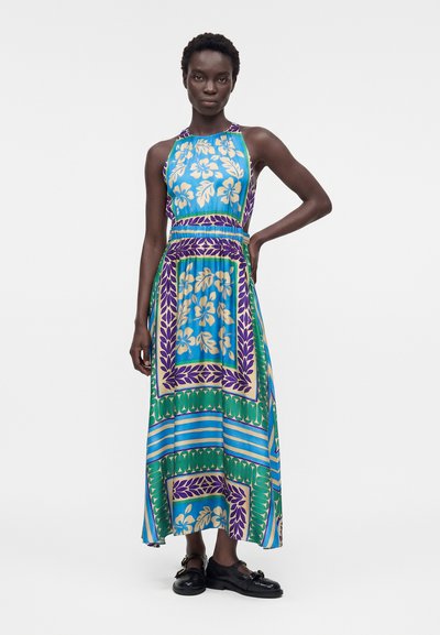 Γυναίκα με μακρύ αμάνικο φόρεμα με μπλε, πράσινα, μοβ και μπεζ ανθισμένα και γεωμετρικά σχέδια, στέκεται με το ένα χέρι στη μέση.