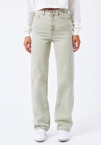 Jeans verde chiaro a vita alta con vestibilità comoda, cuciture in vista e tasche frontali, abbinati a sneaker bianche. Tessuto denim liscio.