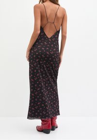 Robe longue fleurie noire avec fines bretelles et dos en V. Le tissu semble lisse, avec de petits motifs floraux rouges et verts. Associée à des bottines rouges à la cheville.