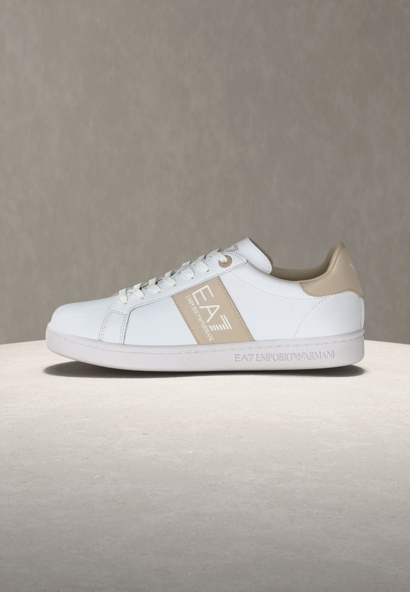 Λευκό αθλητικό παπούτσι χαμηλού προφίλ με μπεζ λεπτομέρειες και λογότυπο EA7 Emporio Armani στο πλάι και στη σόλα, φωτογραφημένο πάνω σε ουδέτερο φόντο.
