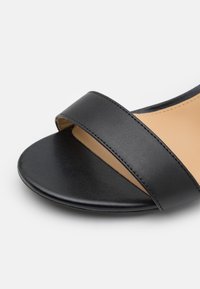 Sandale en cuir noir avec une large sangle sur le dessus et une semelle intérieure lisse beige. La semelle est ronde et élégante, avec des détails de couture subtils.